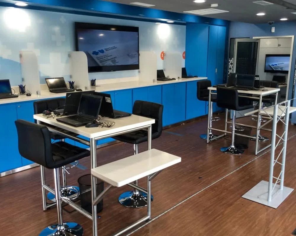 Interior view of a modern Blue Cross Blue Shield mobile command center trailer-2 with blue cabinets, multiple laptop workstations, high chairs, a long screen, and a dark wood-look floor.