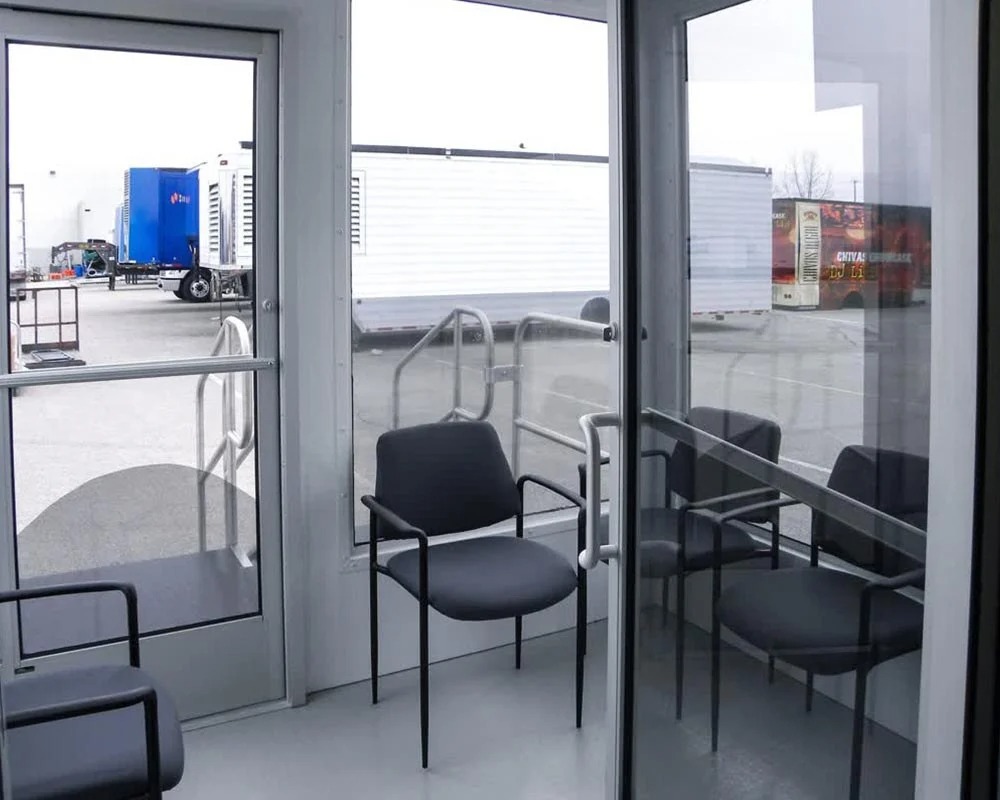 The inside waiting area of a diabetes medical trailer, showing multiple black chairs and a glass door with a view of other specialized medical trailers parked outside.