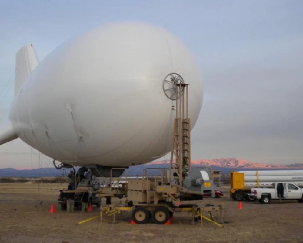 Large white military blimp or aerostat being docked on a specialized mobile fabrication platform in a desert landscape.