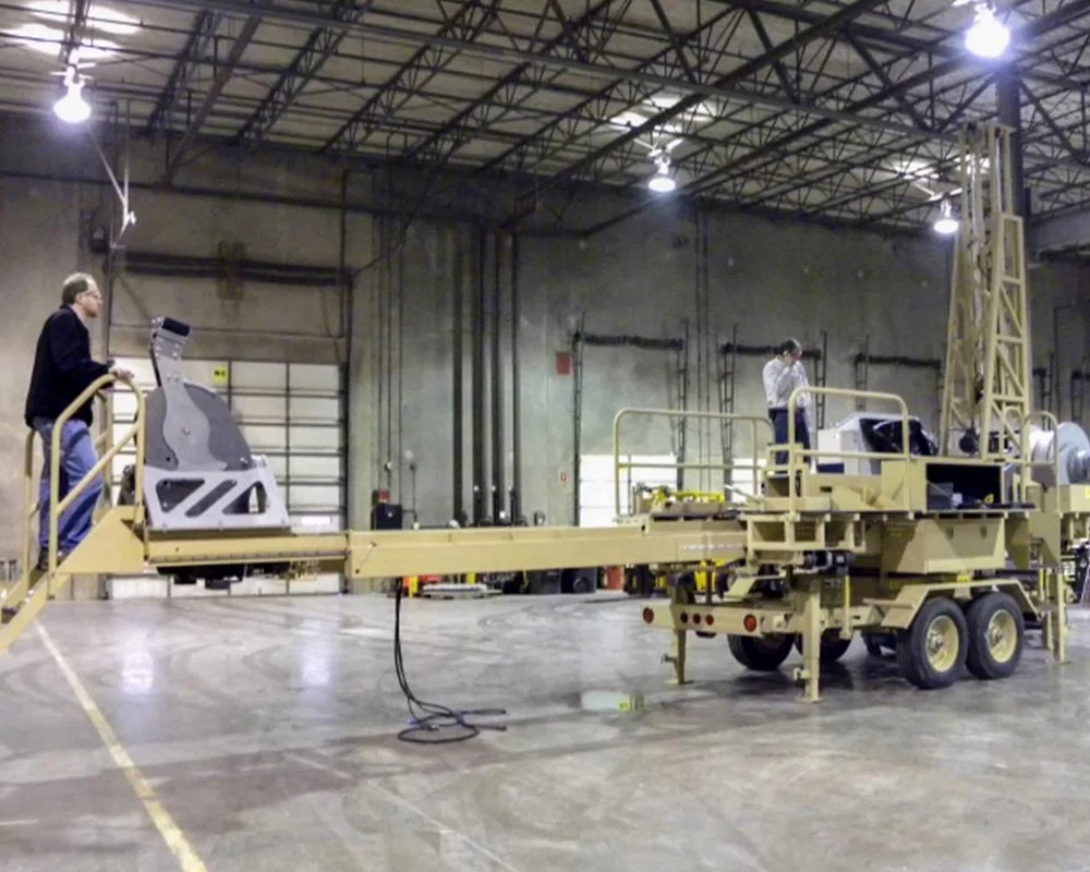 Military fabrication platform with a docking arm and personnel, on a multi-wheeled trailer inside a large maintenance hangar.