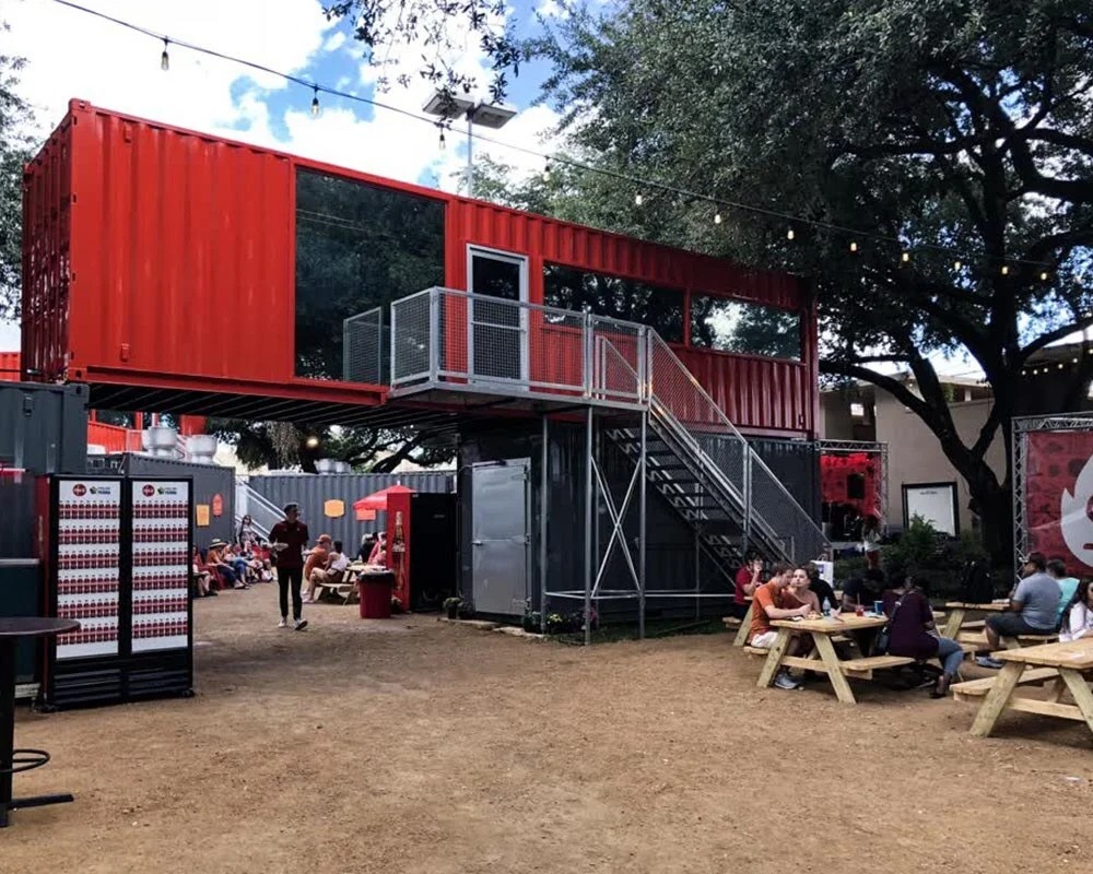 Red sutside shipping container restaurant with an upstairs deck, metal staircase, and outdoor picnic table seating.