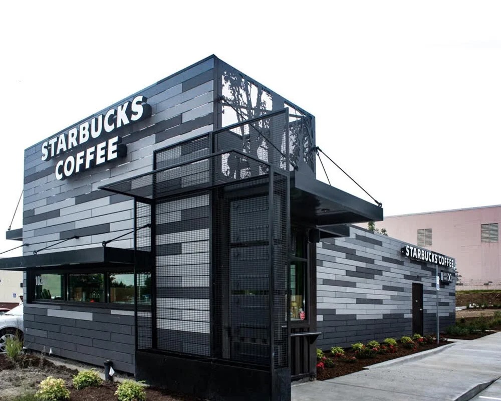 Starbucks Coffee shipping container conversion into a modern drive-through with gray siding, black metal accents, and a small upper viewing deck.