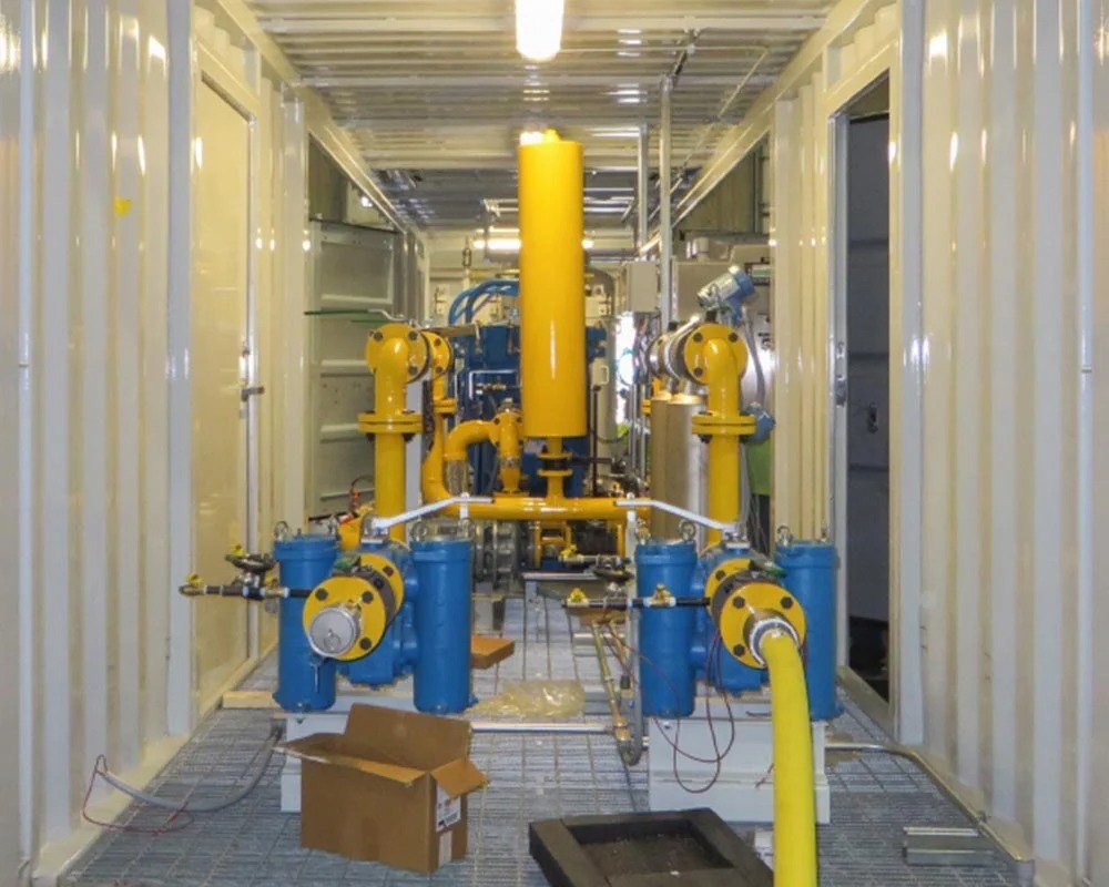 Interior view of a shipping container housing industrial fabrication equipment with blue pumps, yellow piping, and a mesh floor grating.