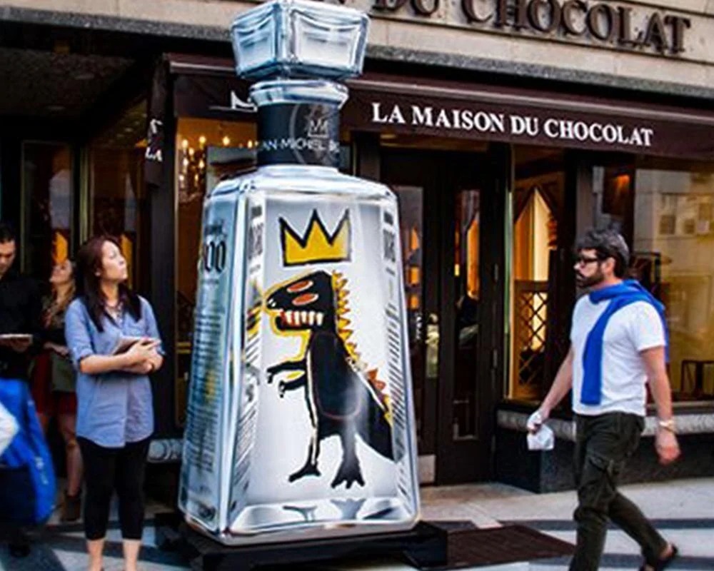 A giant, clear acrylic or glass replica of a 1800 Tequila bottle featuring the artwork of Jean-Michel Basquiat (a black dinosaur with a crown) on display outside a storefront labeled