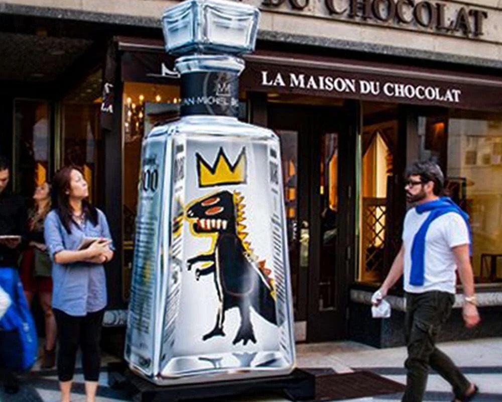 Street photo of a massive, clear 1800 Tequila bottle installation featuring a black and yellow Jean-Michel Basquiat crown and creature artwork, with a man and woman standing by La Maison du Chocolat storefront.