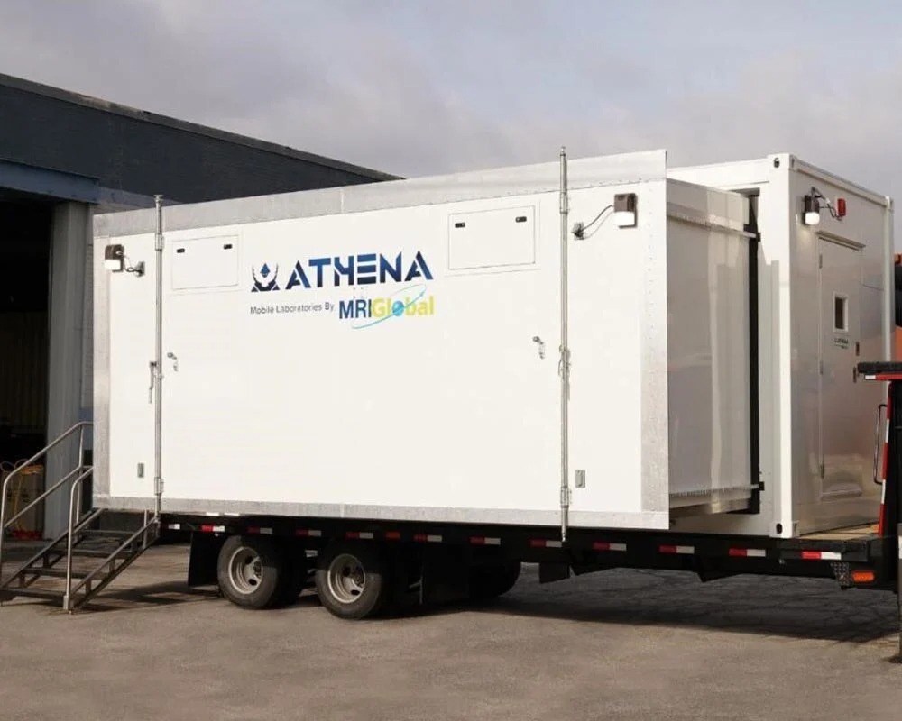A white Athena mobile laboratory unit on a flatbed trailer, with an exterior staircase for access. The transportable lab has the Athena and MRIGlobal logos and is ready for deployment.