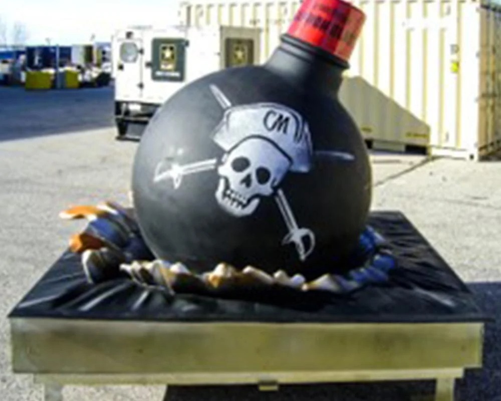 An oversized, sculpted replica of a round, black cartoon bomb with a red and black checkered fuse, painted with a skull and crossbones logo that includes the initials
