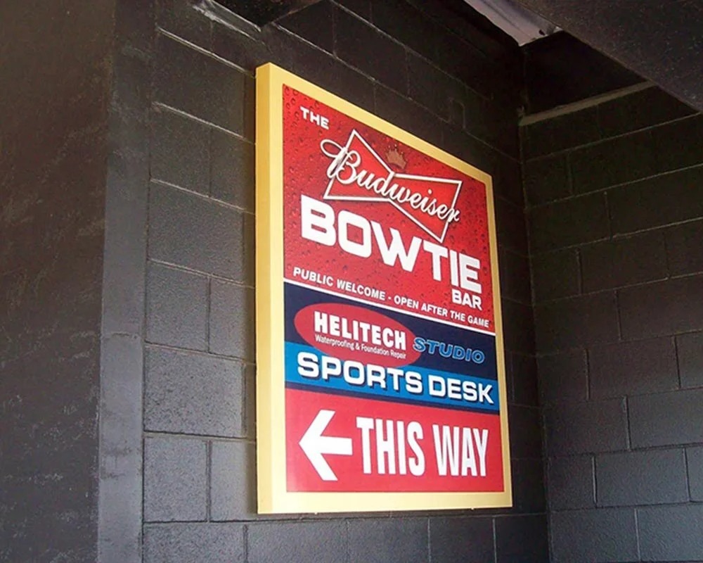 Outdoor backlit box sign for the Budweiser Bowtie Bar and Sports Desk directing visitors with a large white arrow.