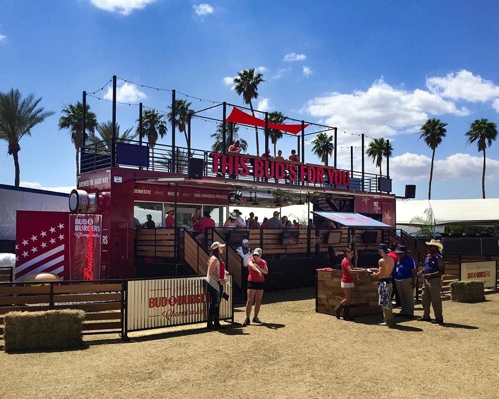 Bud and Burgers two-story mobile marketing trailer at an outdoor festival surrounded by people and palm trees.