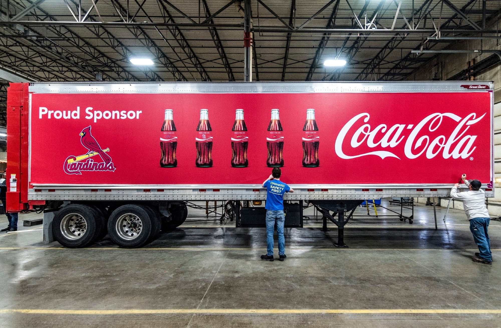 Coca-Cola semi-trailer truck with a bright red wrap featuring the Cardinals logo, six glass Coke bottles, and the text "Proud Sponsor" and "Coca-Cola" being applied by workers in a warehouse.