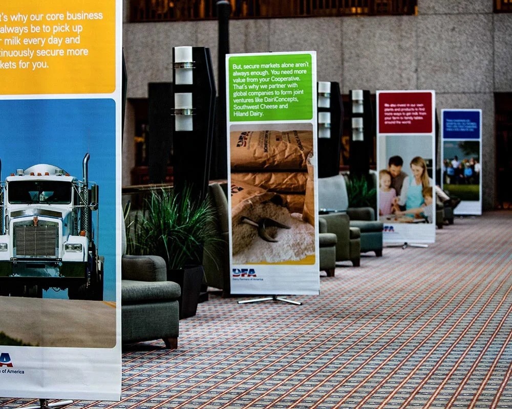 Indoor trade show environment with DFA branded vertical banner stands displaying dairy products and cooperative mission text.
