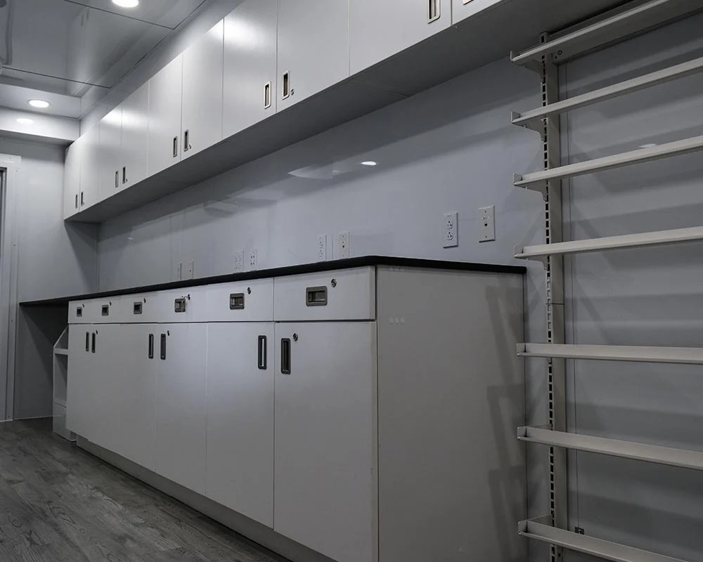 The interior of a Hy-Vee Mobile Lab Trailer, showcasing a long counter with built-in storage cabinets and electrical outlets. The on-site lab also includes adjustable shelving and overhead cabinets for supplies.