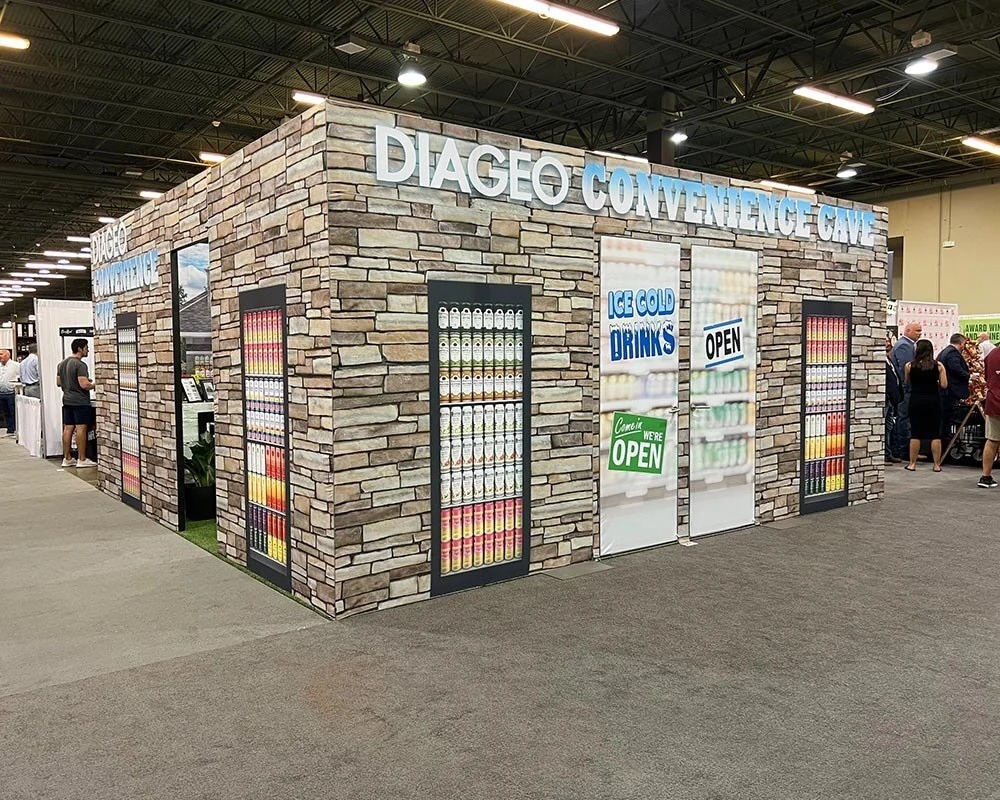 Diageo Convenience Cave booth at a trade show, designed to look like a stone building with branded coolers displaying various canned beverages, and signs for