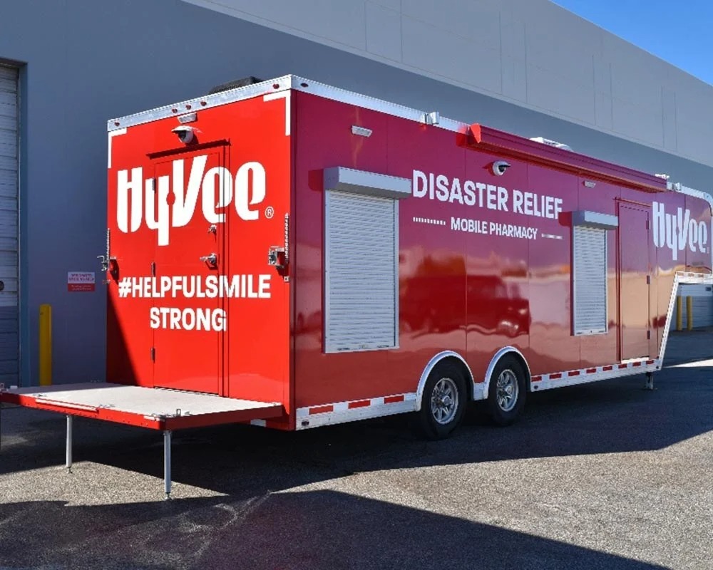 A large red Hy-Vee Disaster Relief Mobile Pharmacy trailer. The side features roll-down service windows and the rear has a fold-down platform, indicating its use for providing quick-response on-site pharmacy services.