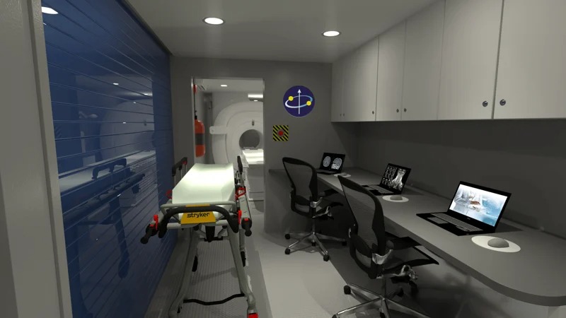 The interior of a specialty medical trailer designed for advanced imaging. The space includes a rolling stretcher, a work desk with computer monitors showing brain scans, and a large MRI machine visible in the back. This mobile imaging unit is equipped for complex diagnostics.
