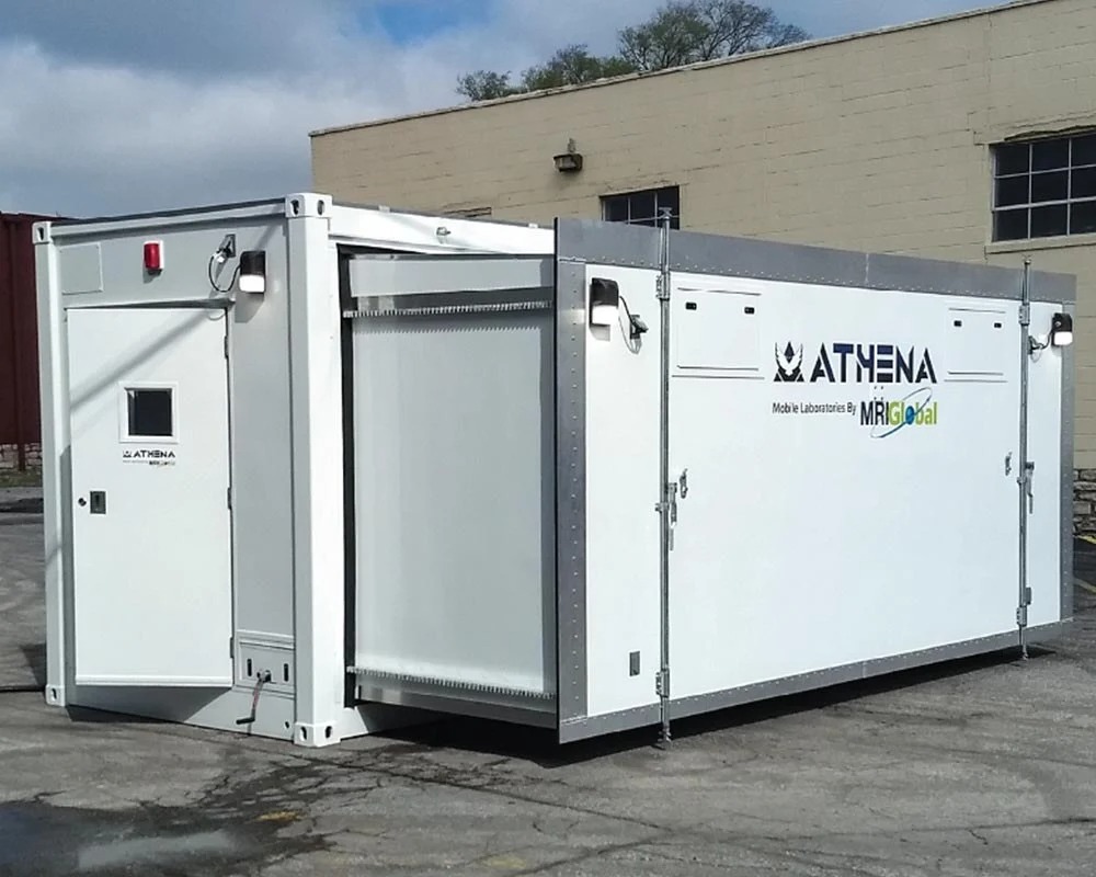 A modern Athena mobile laboratory unit with a slide-out section extended, showing its expandable interior. The portable lab container has the MRIGlobal and Athena logos, indicating its use for on-site medical and scientific testing.
