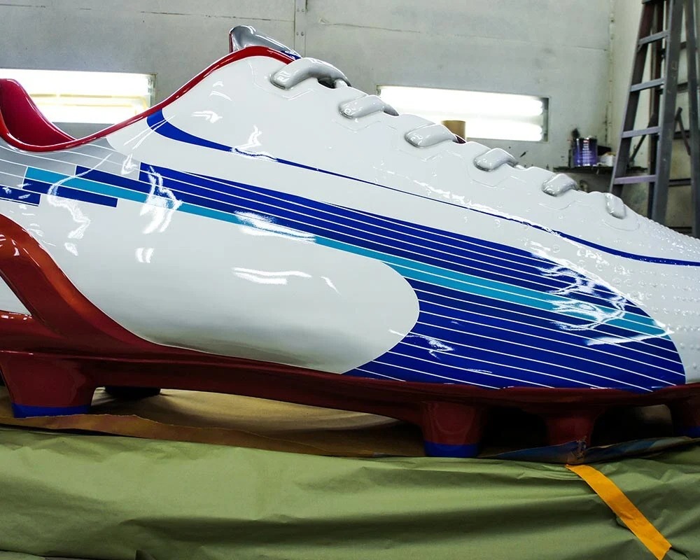 Close-up of a massive, glossy, white fiberglass or molded replica of a Puma soccer cleat, featuring blue, gray, and red graphics, white laces, and mounted on a red base designed to look like a set of cleats, likely for a promotional display.