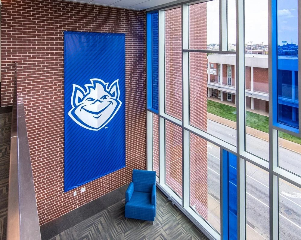 Indoor St. Louis University (SLU) Billiken mascot banner hanging on a brick wall next to large windows.