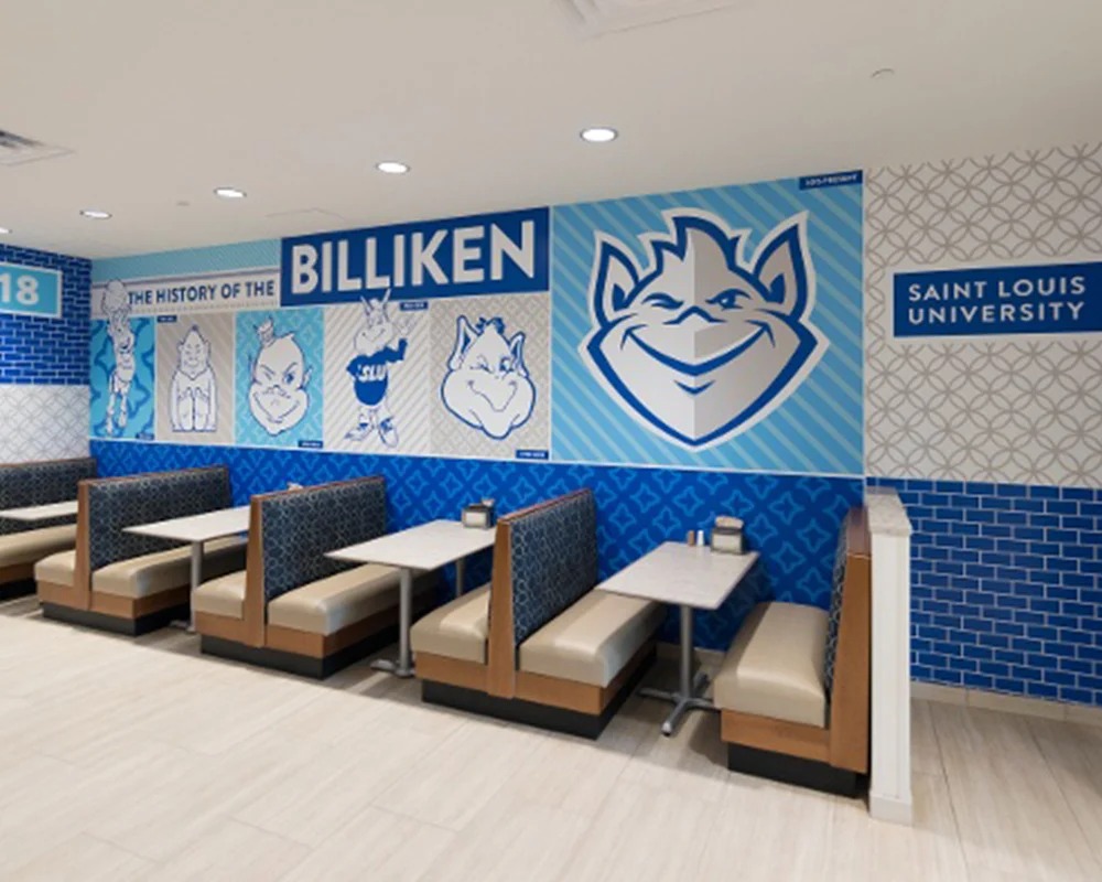 Interior view of a modern dining area or student center at Saint Louis University (SLU), featuring rows of booths and a large blue and white wall graphic timeline titled