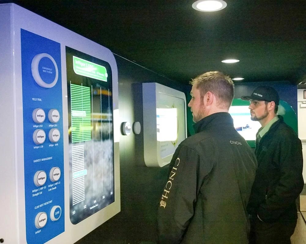 Two men viewing an interactive digital display panel inside a custom agricultural technology showroom trailer.