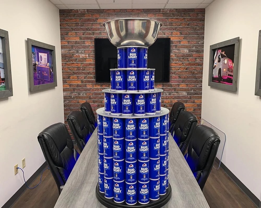 Trophy crafted from dozens of blue Bud Light beer cans topped with a silver bowl, centerpiece on a gray conference table with black leather chairs and a brick wall backdrop.