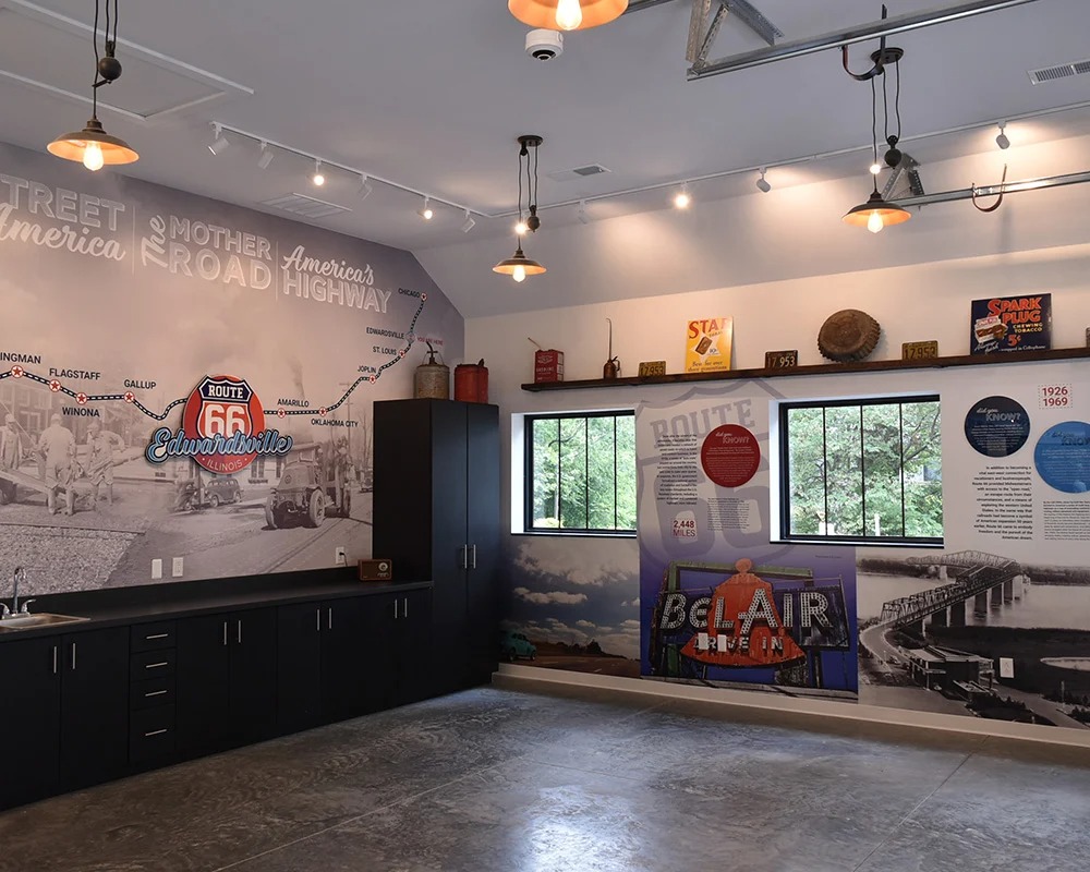 Interior view of the West End Station Route 66 Museum in Edwardsville, Illinois, featuring large wall graphics. The graphics include a Route 66 map stretching from Chicago to Los Angeles, a vintage image of road construction, and a timeline section with a photo of the Chain of Rocks Bridge and a retro Bel-Air Motel neon sign graphic.