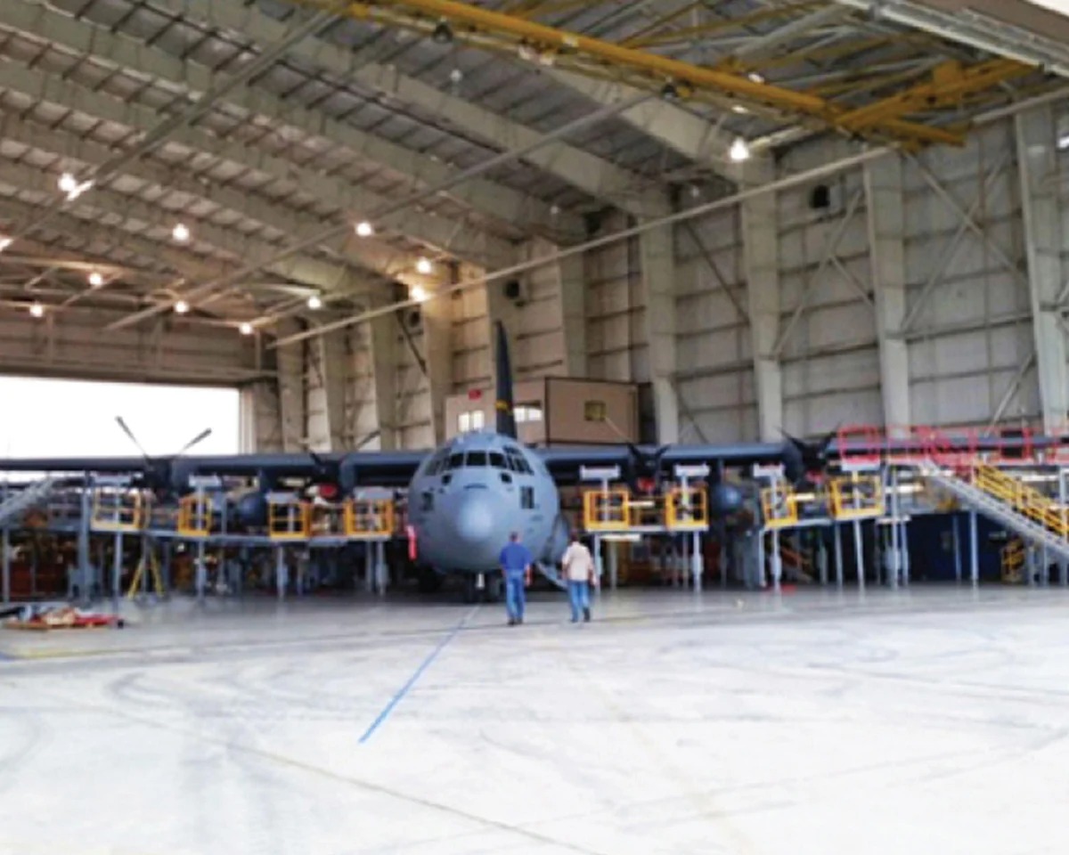 Military fabrication hangar showing a large C-130 Hercules aircraft surrounded by custom scaffolding and maintenance platforms.