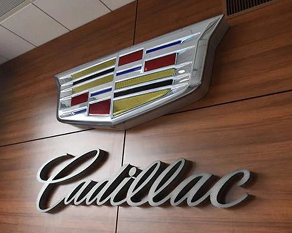 Interior Cadillac dealership sign featuring dimensional chrome logo crest and metallic script lettering on a wood panel wall.