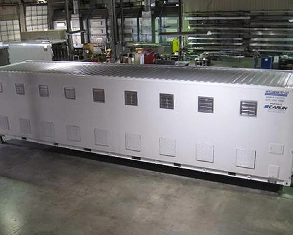 Exterior view of a long, white Carlin commercial mobile kitchen trailer or container, showing multiple small windows, ventilation panels, and its large size inside a warehouse or factory.