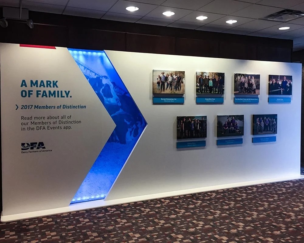 Dairy Farmers of America (DFA) 2017 Members of Distinction backlit display wall with an illuminated blue arrow graphic and seven framed photos of honoree families.
