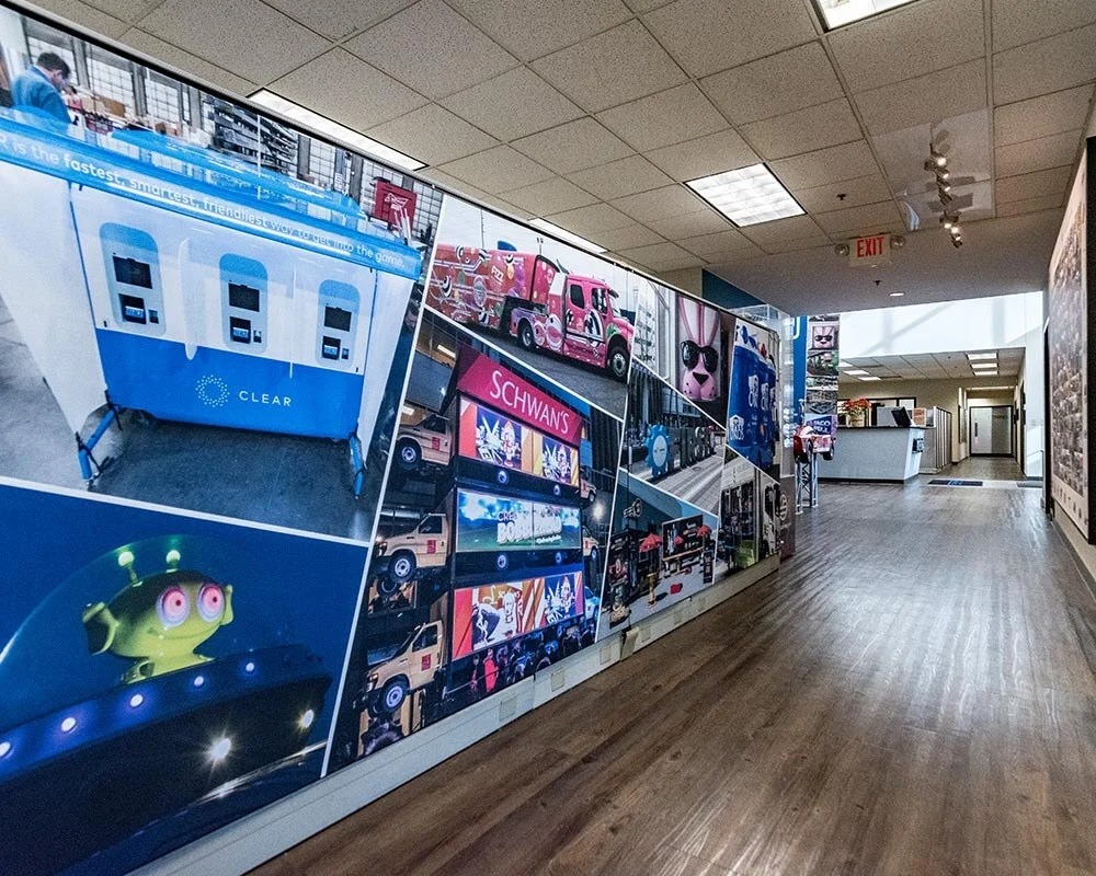 Long office corridor wall covered in a collage-style mural of various experiential graphics, including a blue 'CLEAR' branded container, Schwan's truck wraps, and an underwater alien character display.