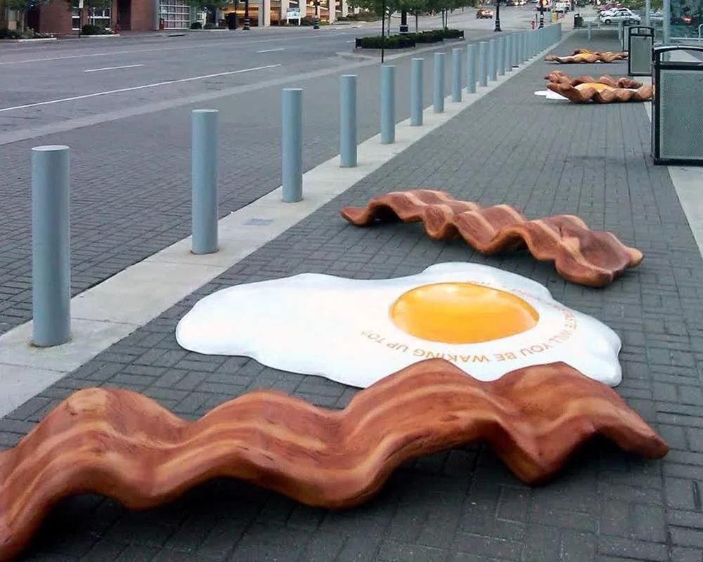 Experiential marketing display featuring oversized, sculptural replicas of a sunny-side-up egg and several wavy strips of bacon, scattered across a brick sidewalk next to a street lined with bollards.