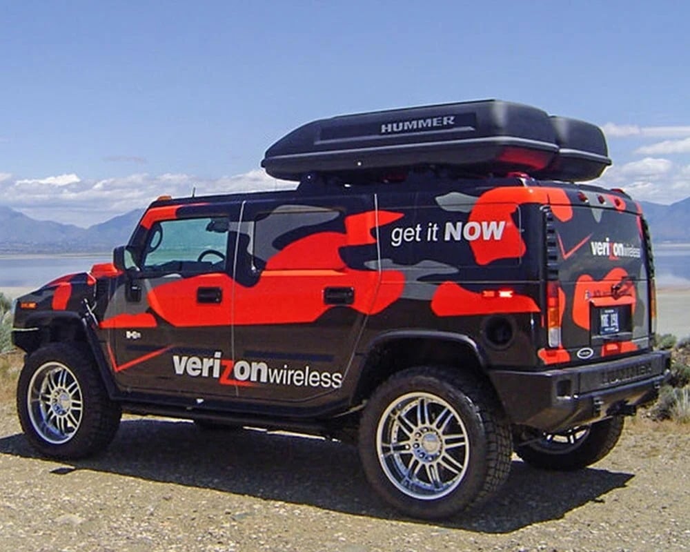 Black Hummer H2 SUV with a distinctive custom vinyl vehicle wrap in red, gray, and black camouflage, promoting Verizon Wireless with the text