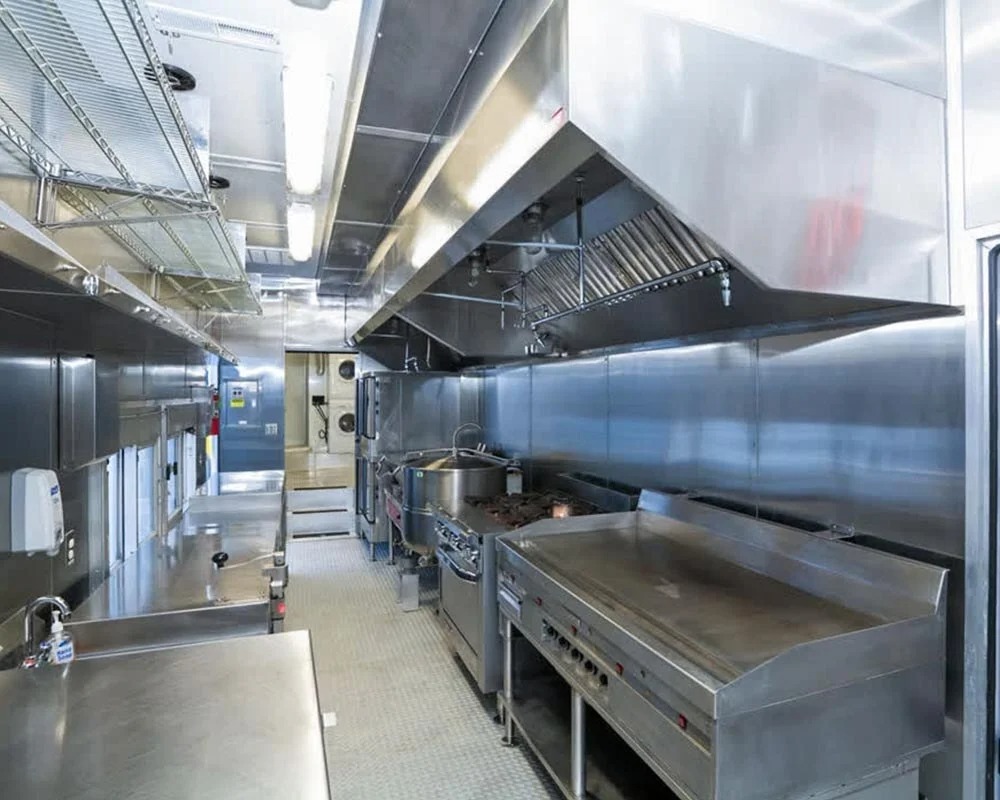 Interior view of a professional commercial mobile kitchen trailer showing a large flat grill or griddle, gas range, massive exhaust hood, and stainless steel prep counters.