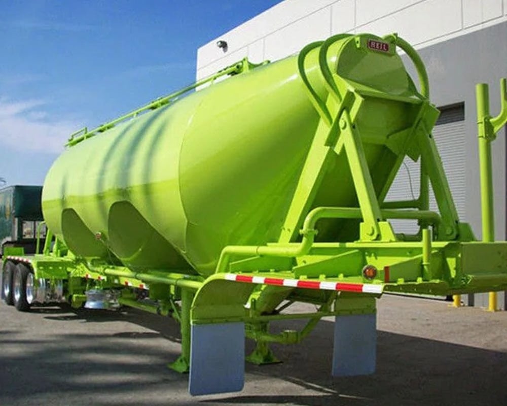 Close-up of the rear of a large industrial dry bulk pneumatic tank trailer, painted a vibrant, bright lime green, with 'HEIL' branding visible on the frame, featuring two mud flaps and parked against a light-colored commercial building.