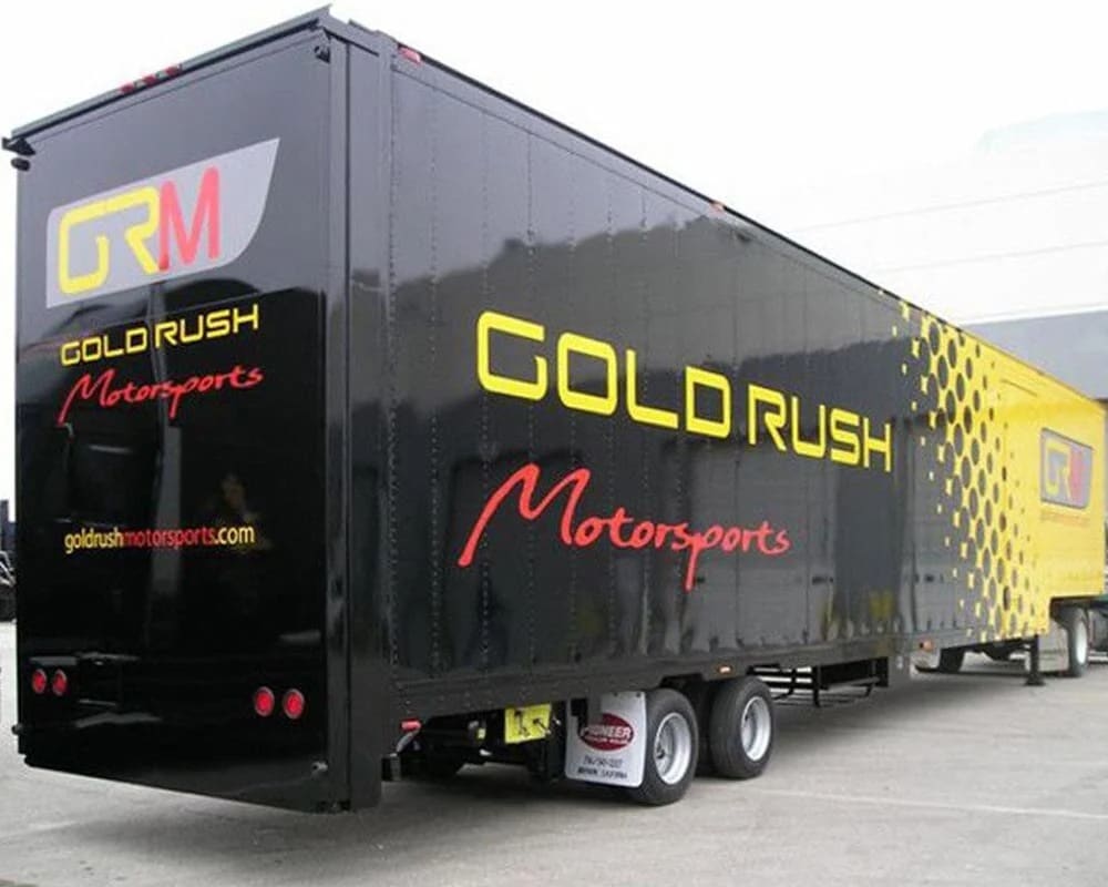 Rear three-quarter view of a black enclosed semi-trailer for GOLD RUSH Motorsports, featuring large yellow and red branding, a yellow honeycomb/dot pattern graphic on the side, and the website 'goldrushmotorsports.com'.