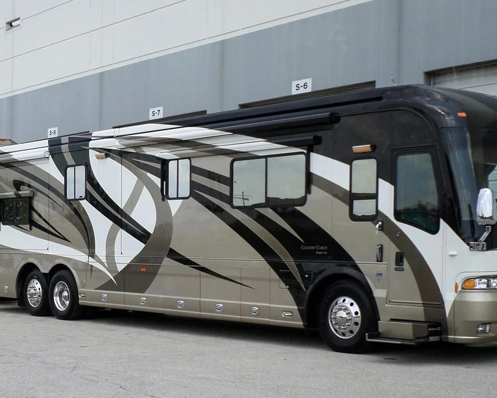 High-end Country Coach motorhome with a distinctive custom paint and graphic design in black, white, and tan, parked outside a warehouse with bay doors labeled S-6, S-7, and S-8.