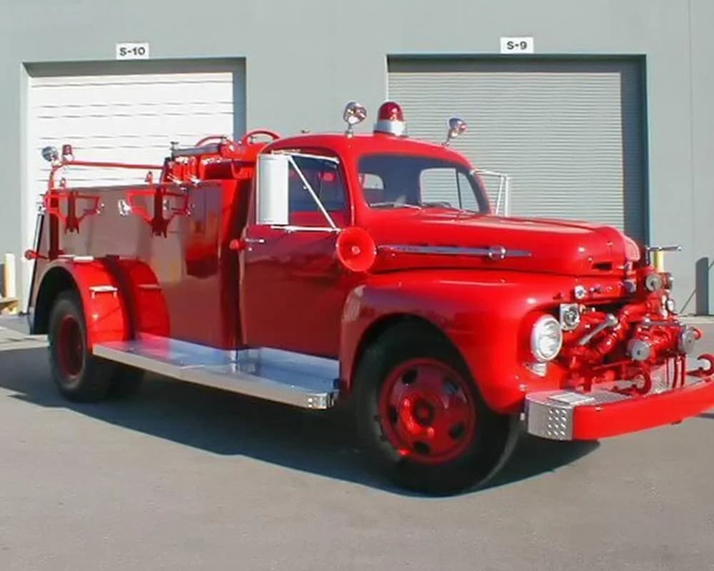 Fully restored vintage bright red fire truck, likely a classic Ford model, with chrome accents and a single beacon light, parked in front of two gray warehouse bay doors labeled S-10 and S-9.
