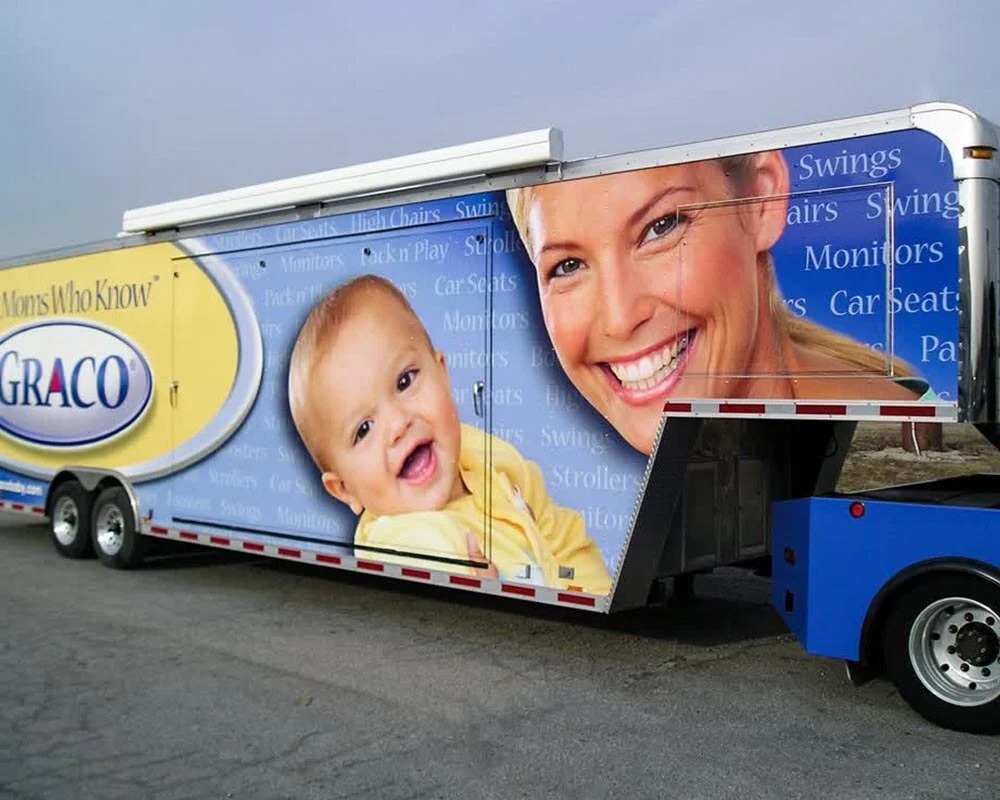 Graco mobile billboard trailer with a large graphic of a smiling baby and mom, featuring text about car seats, swings, monitors, and strollers.