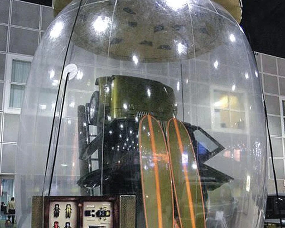 An artistic installation featuring a vintage military Jeep or utility vehicle and a pair of tall wooden skis encased entirely within a massive, clear, jar-shaped inflatable or acrylic dome, displayed indoors.
