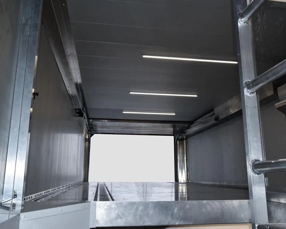 Interior storage motorcycle trailer with shiny aluminum walls, fluorescent strip lighting, and wheel chocks on the floor.