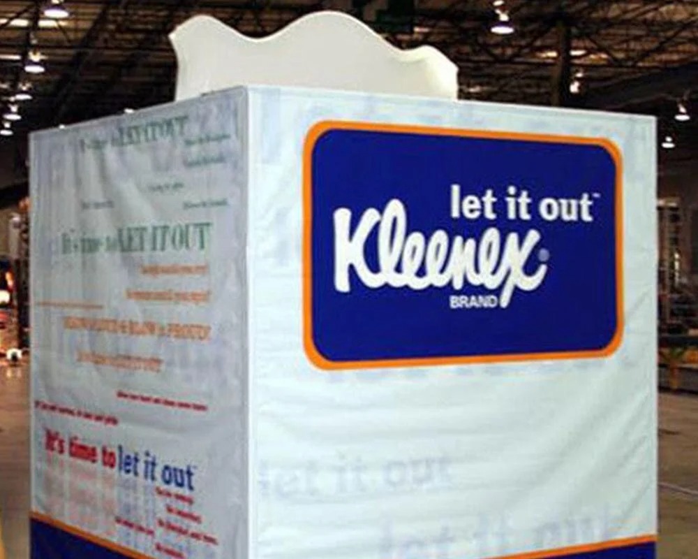 A large, cube-shaped, experiential marketing element designed to look like a giant Kleenex tissue box, featuring the Kleenex logo in a blue and orange border, the slogan