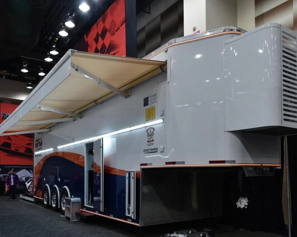 Marco Polo Motorsports transporter trailer exterior with awning extended, featuring blue/orange KTM and MPM branding at an indoor event.