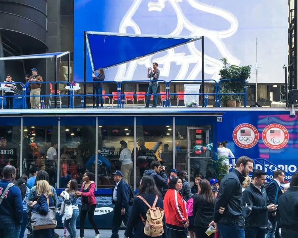 Olympic Team USA two-story mobile showroom trailer with a rooftop deck and large blue graphic in a crowded city square.
