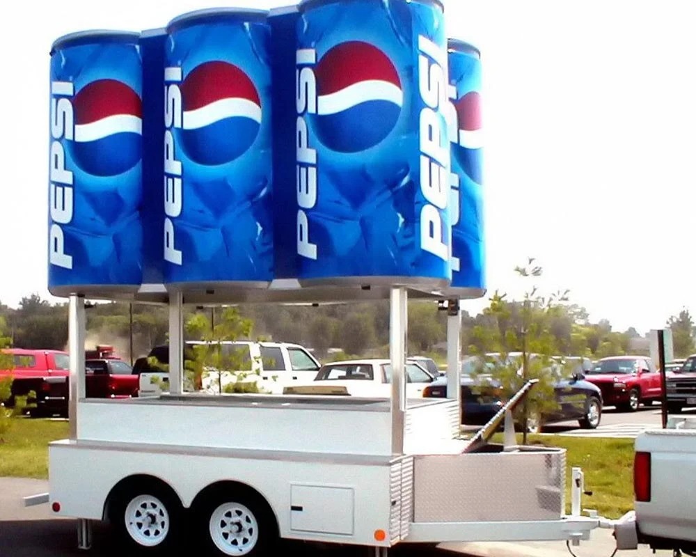 A white, dual-axle concession or promotional trailer featuring an overhead display of four massive, cylindrical replicas of Pepsi soda cans with the iconic blue, white, and red logo, parked in an outdoor setting with other vehicles.