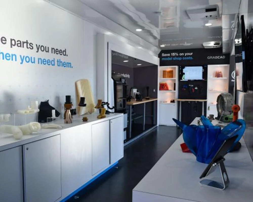 Interior of the Stratasys and GrabCAD mobile showroom with displayed 3D-printed parts and white product cabinets.