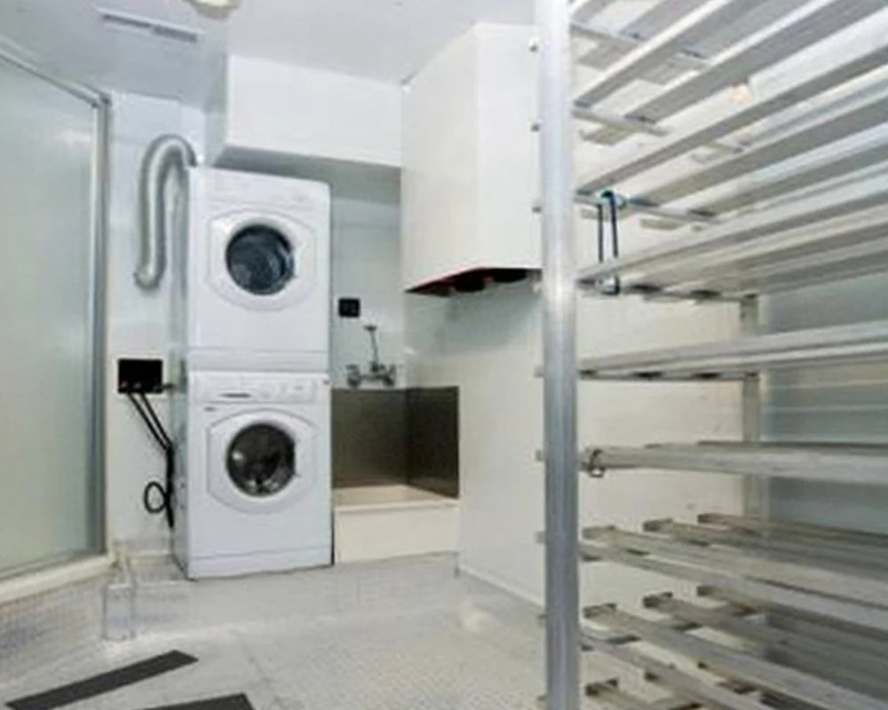 Interior view of a commercial mobile kitchen trailer section converted into a laundry and utility area, featuring a stacked washing machine and dryer unit, and metal storage shelving.
